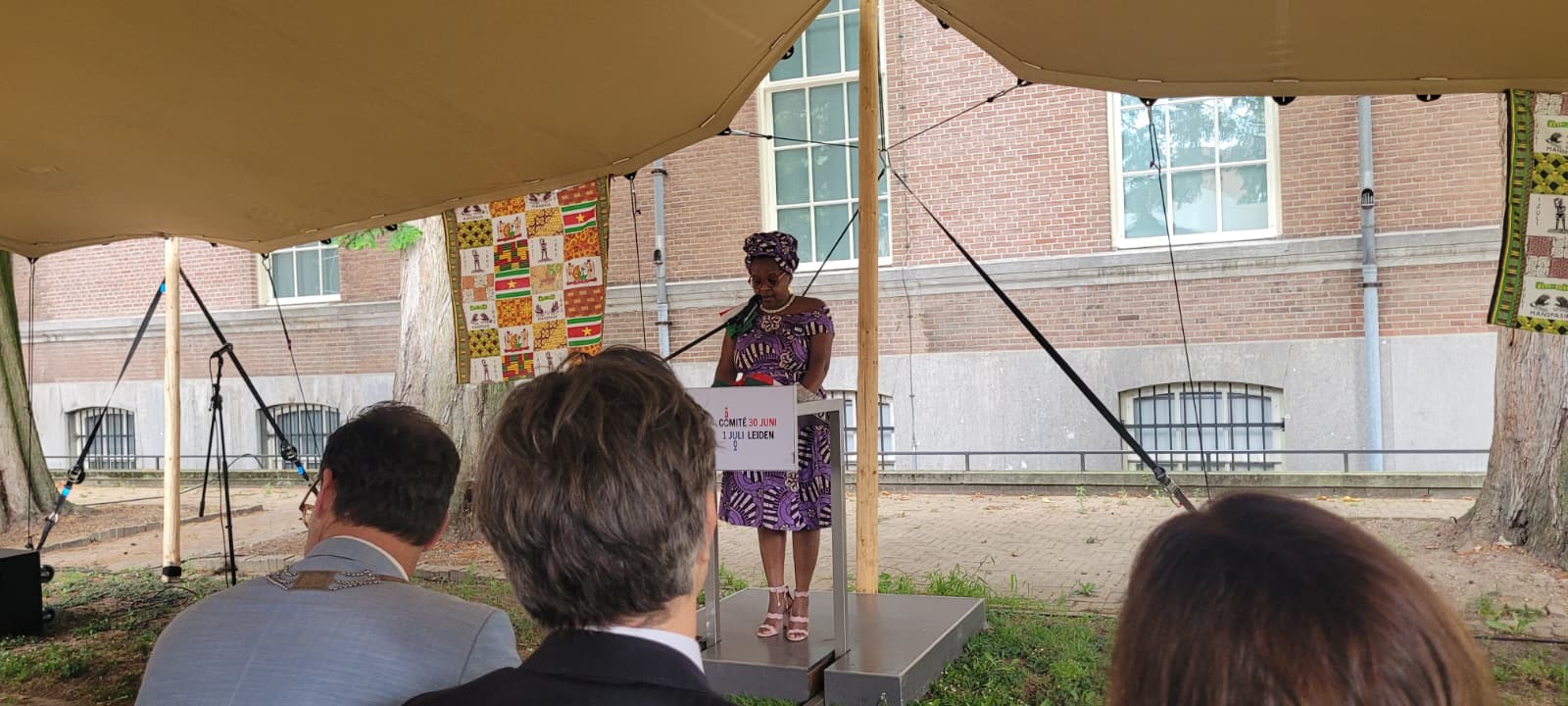 En vrouw in een paarse jurk met traditioneel Surinaamse hoofdoek geeft een toespraak